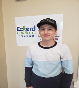 [Image description: A white male with a black hat and a white, blue, and black long-sleeve shirt standing in front of a Eckerds Connect sign on a beige wall.]